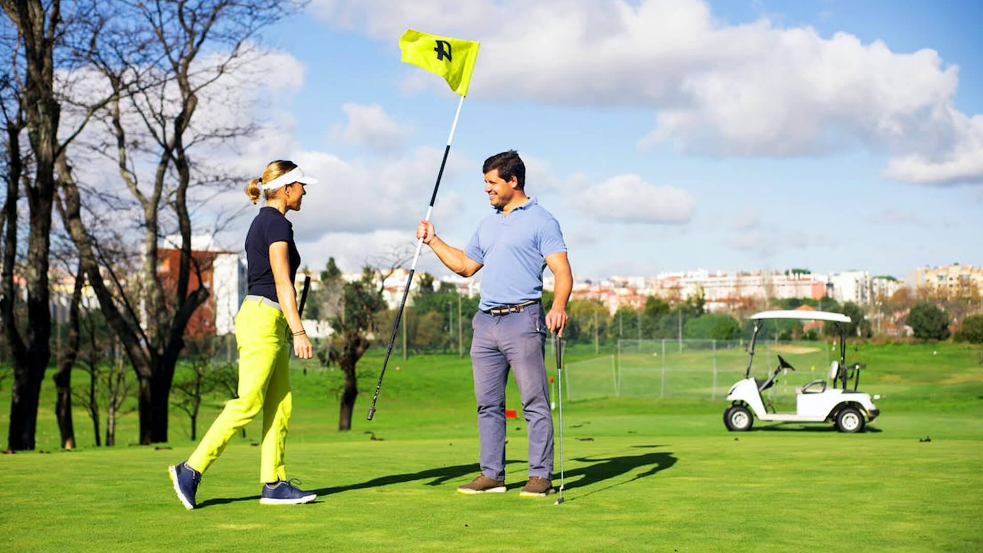 A man and a woman on a golf green. Man is holding the flag.  Golf cart in the background.