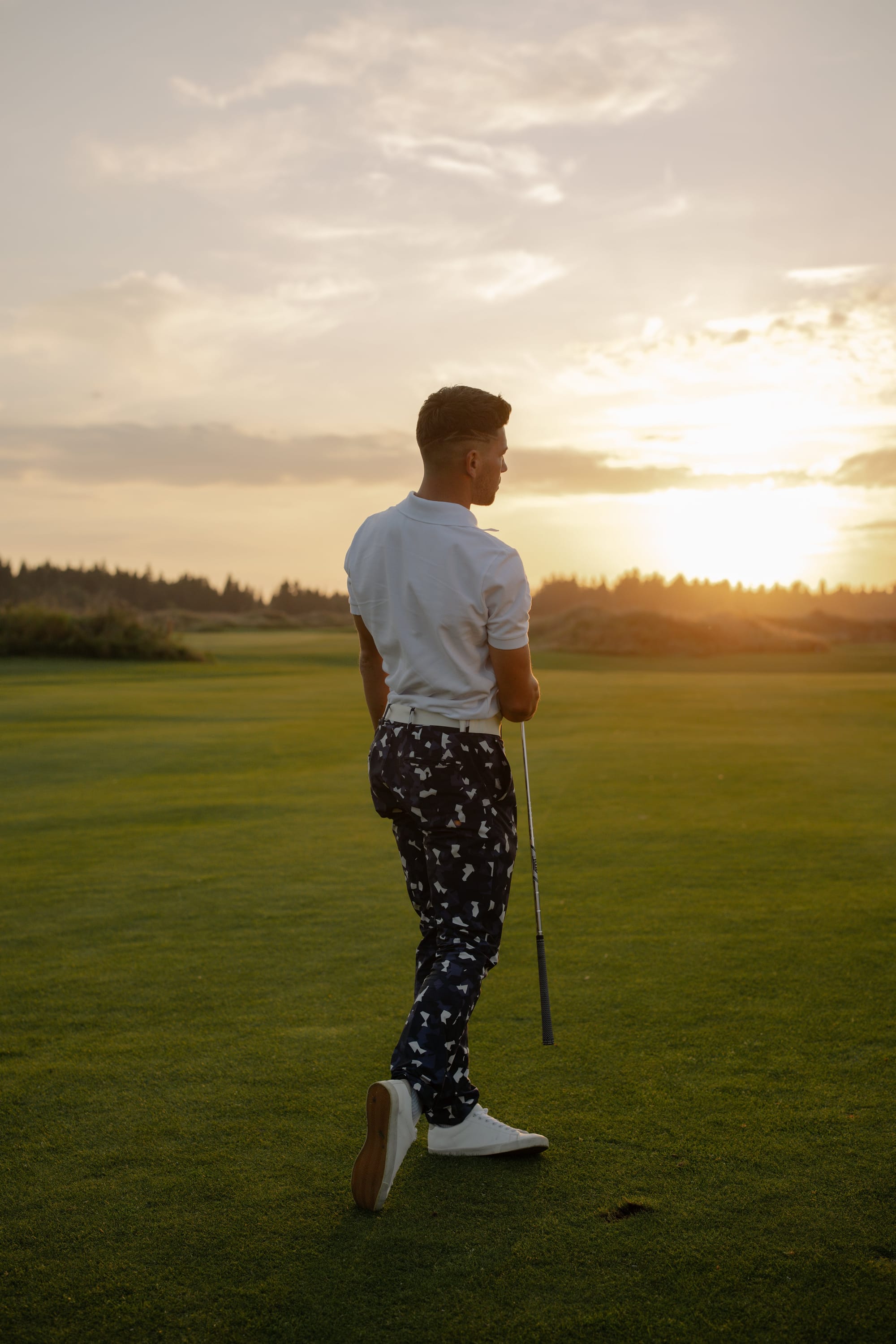 A male golfer holding a golf club and looking out towards a setting sun