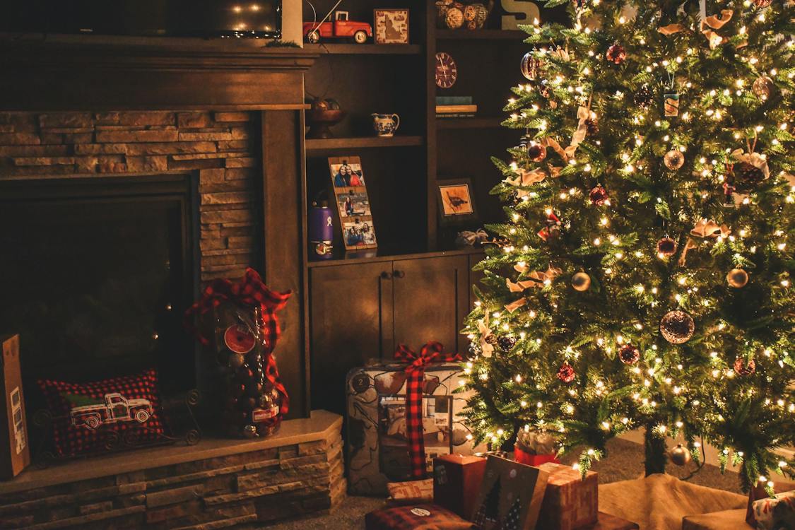 A warm, fully decorated and lit Christmas tree with presents beneath next to a fireplace